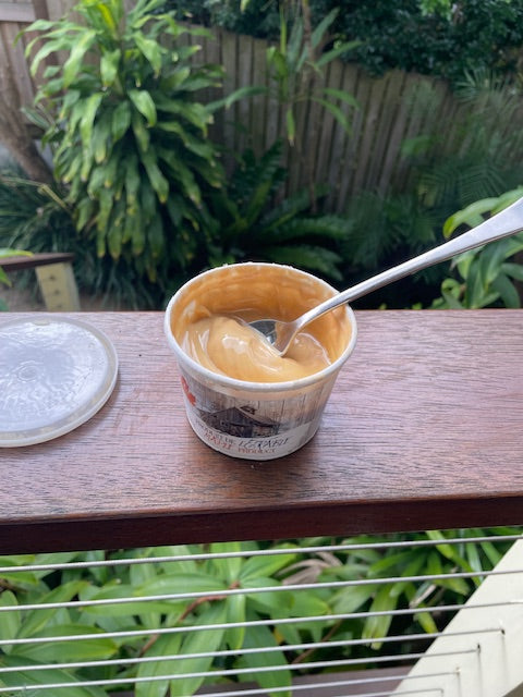 A spoon dipping into an open tub of Canadian maple butter on a wooden deck surrounded by lush greenery