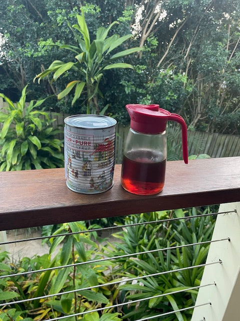 A clear glass pitcher with a red lid and handle, partially filled with a red drink, sits on a wooden balcony railing next to a can of coffee, with lush green tropical foliage in the background.