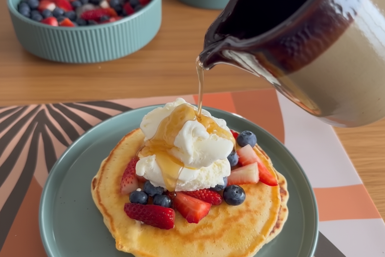 Maple syrup being poured over fluffy pancakes with fresh berries and whipped cream